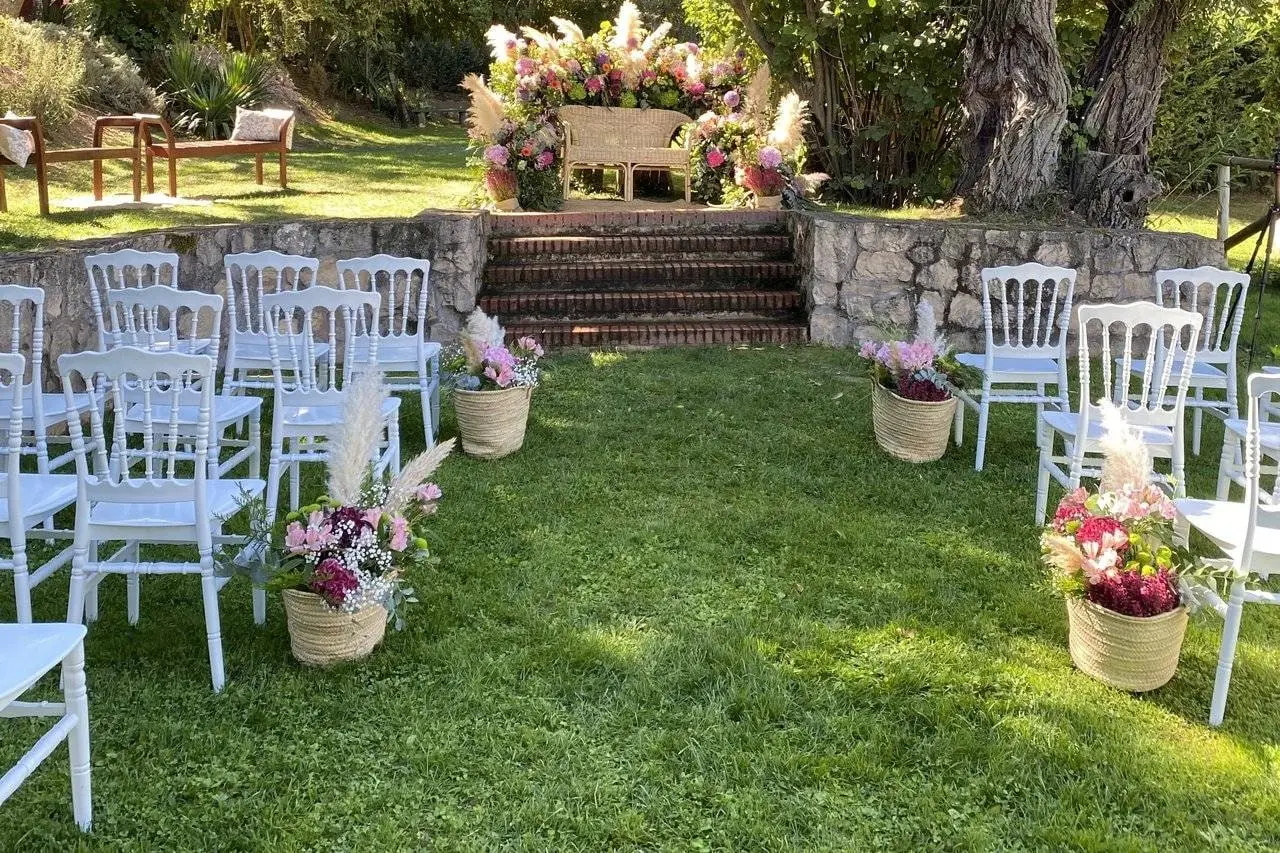 Sillas blancas sobre el césped, cestas de flores a los lados y un arco de flores al fondo, preparadas para una ceremonia