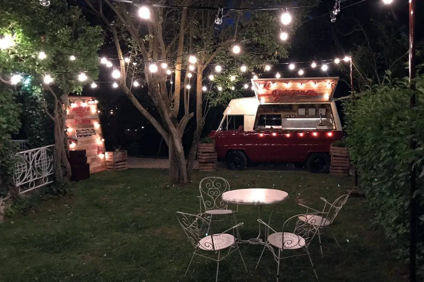 El jardín de noche, con guirnaldas de bombillas entre los árboles, una furgoneta roja y una mesa con sillas de hierro