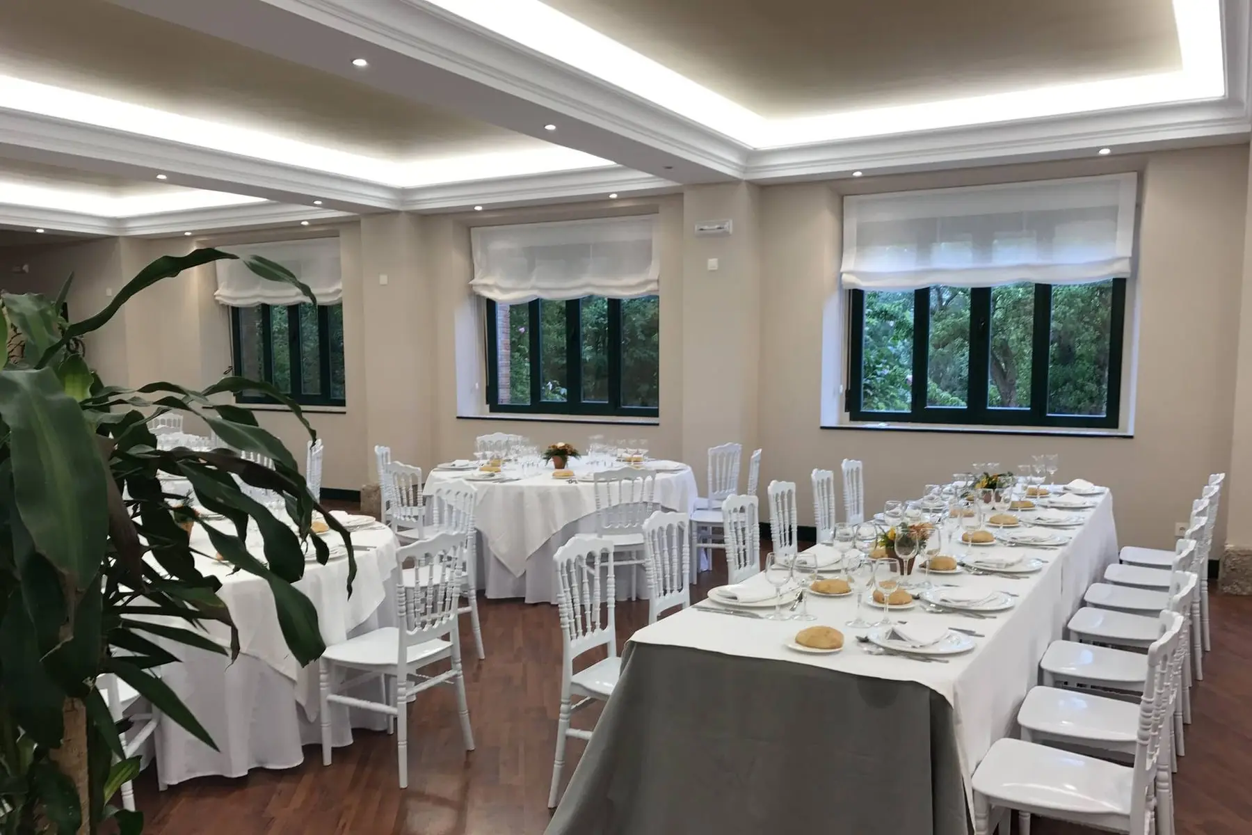 El salón con mesas redondas y una larga mesa imperial vestidas de blanco, sillas blancas y ventanas al jardín