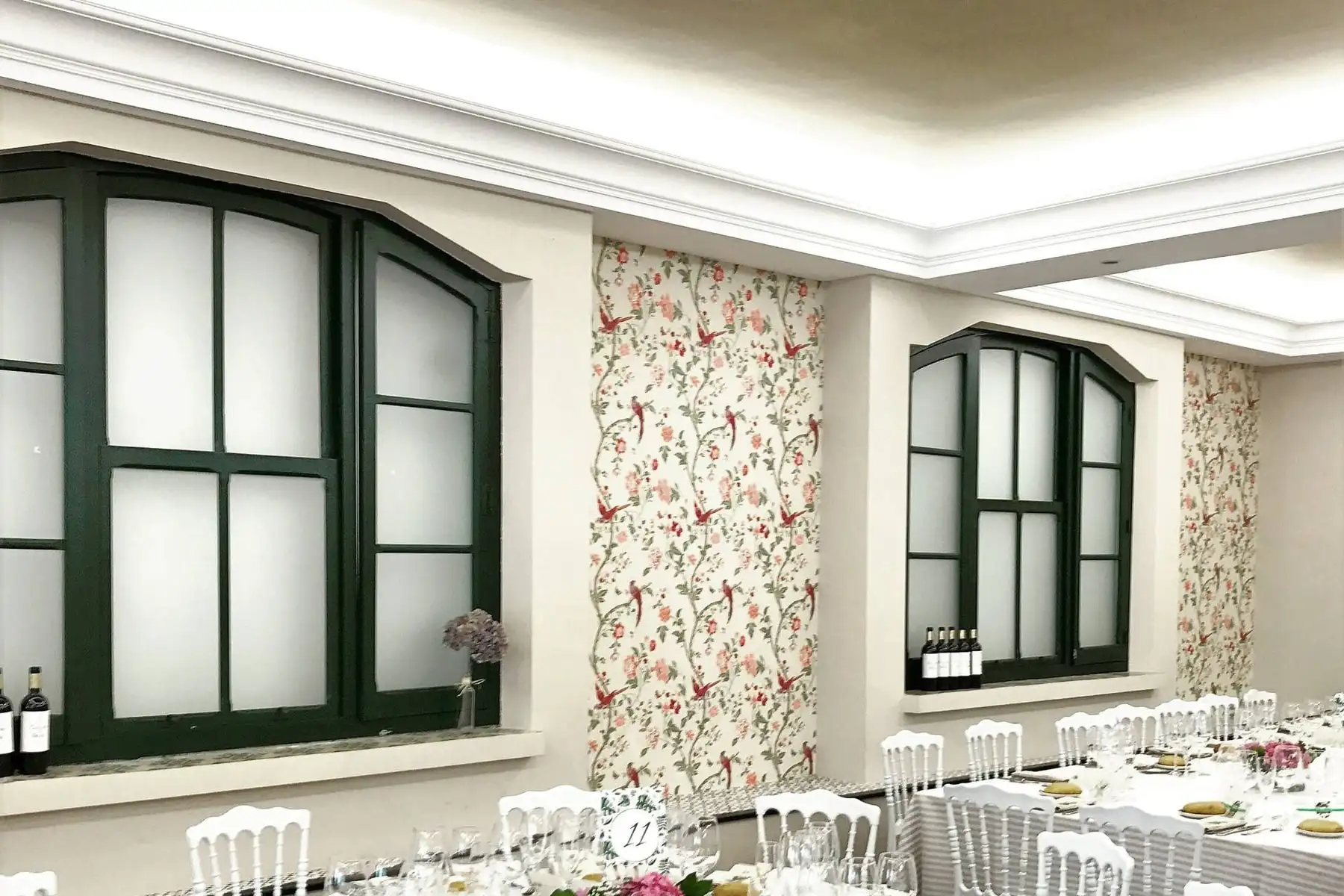 Una esquina del salón con ventanales de marcos verdes, papel pintado de flores y aves, y las mesas del banquete